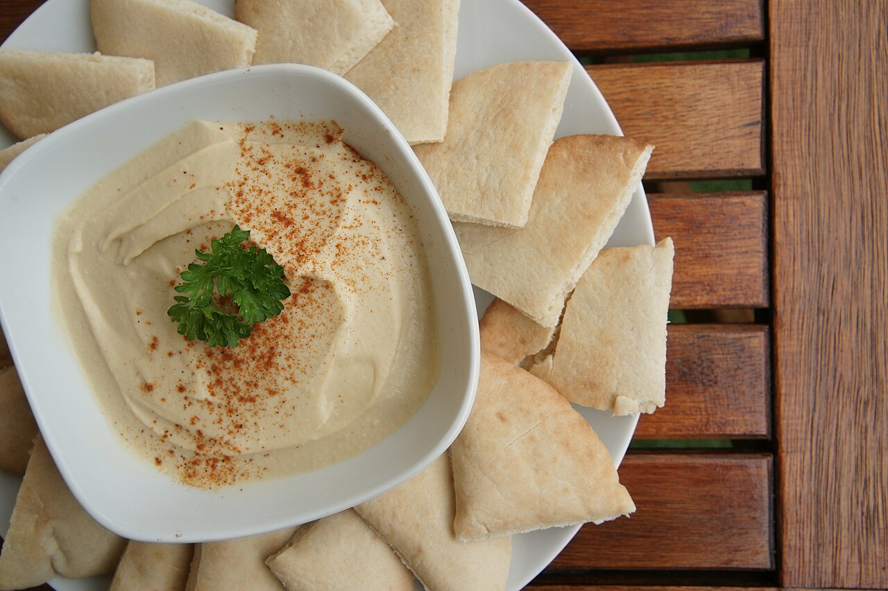 A bowl of hummus garnished with paprika and herbs beside pita bread, illustrative of a pita and hummus snack tray